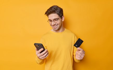 Cheerful guy with dark hair uses mobile phone and banking card for making shopping online orders something in online store enters information does secure payment isolated over yellow background