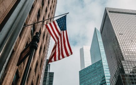 the USA or United States of America flag on a flagpole near skyscrapers under a cloudy sky