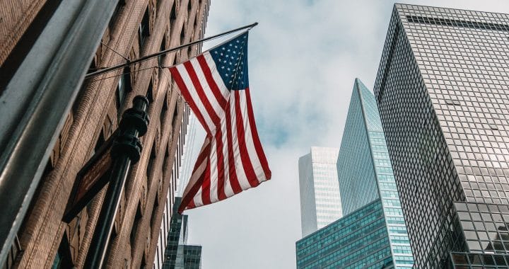 the USA or United States of America flag on a flagpole near skyscrapers under a cloudy sky